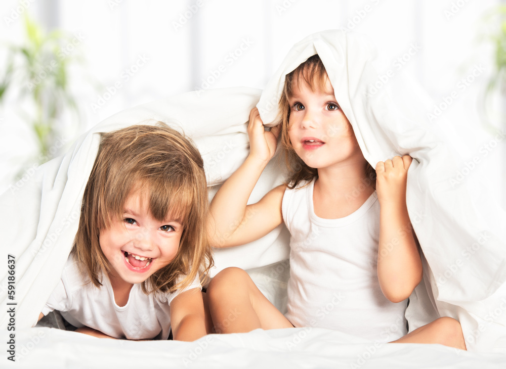 快乐的小女孩双胞胎妹妹躺在床上的毯子下