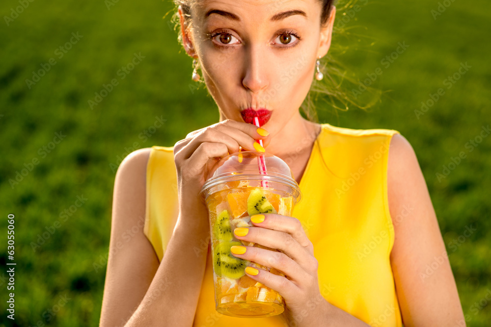 年轻女子在绿色gr上喝透明杯子里的水果