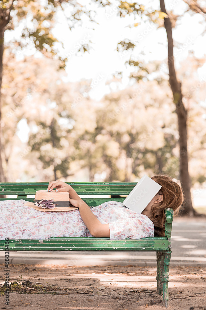 女孩睡在公园的椅子上，脸上戴着书，戴着帽子