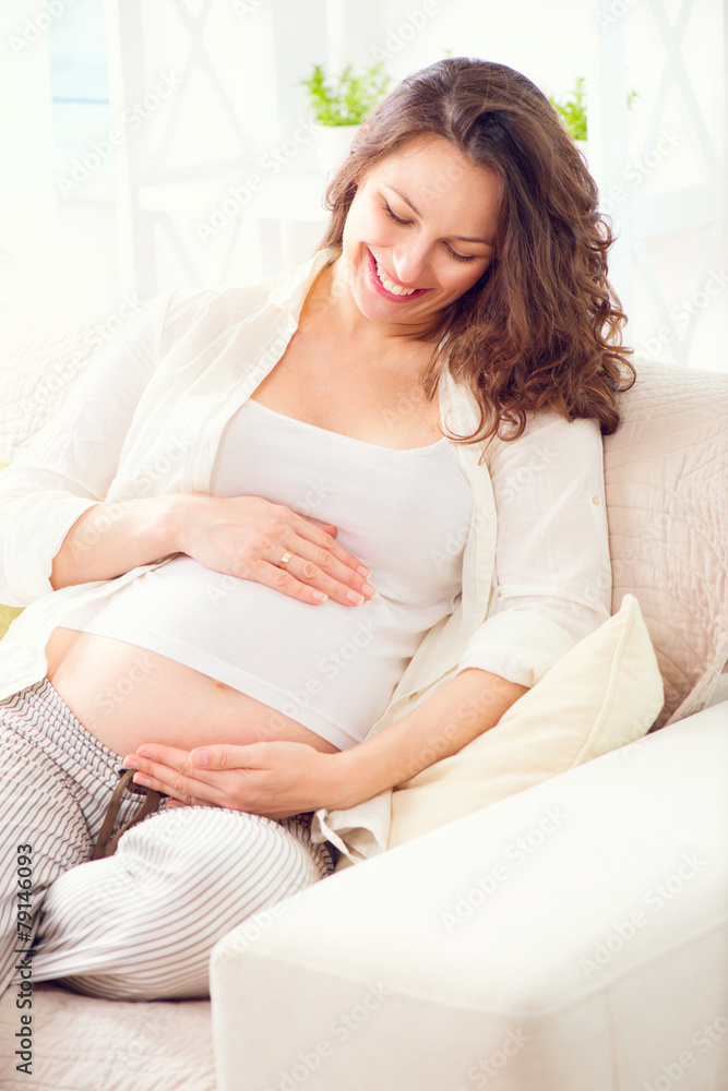 怀孕快乐的女人坐在沙发上抚摸她的肚子