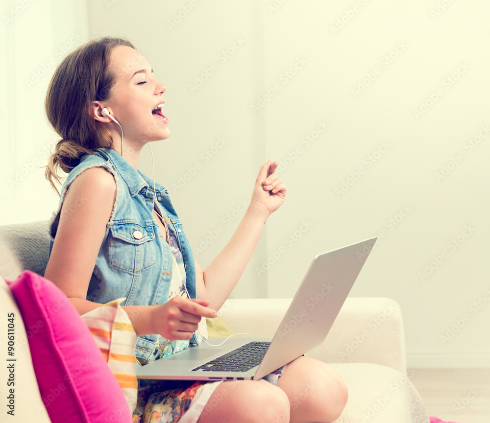 漂亮的十几岁女孩坐在家里的沙发上，用笔记本电脑。用耳机欣赏音乐