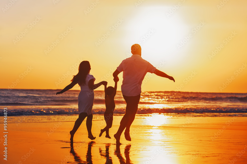 Happy family - father, mother, baby son hold hands and run with fun along edge of sunset sea on blac