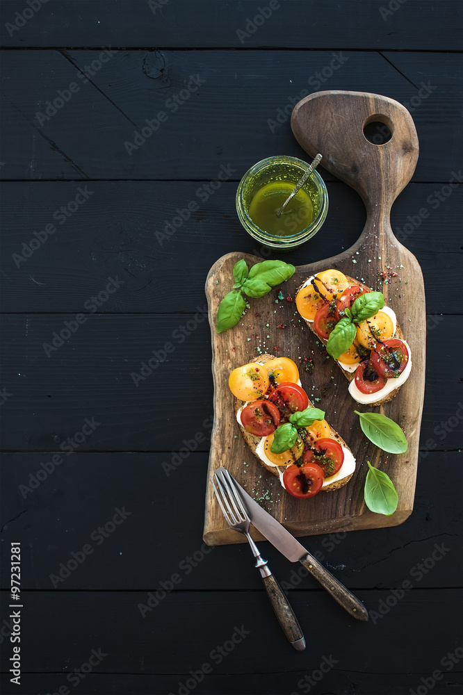 深色木制砧板上的番茄、马苏里拉奶酪和罗勒三明治、香蒜酱罐、餐具