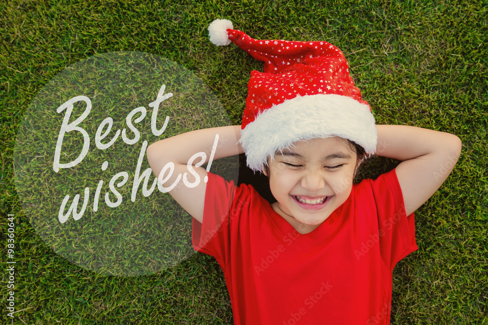 绿色草地上戴着圣诞帽的亚洲小女孩，带着最美好的祝愿
