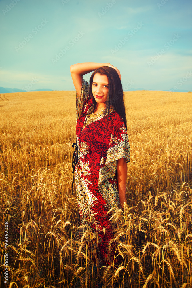 微笑的年轻女子，穿着装饰性连衣裙，站在日落的麦田上。自然背景。