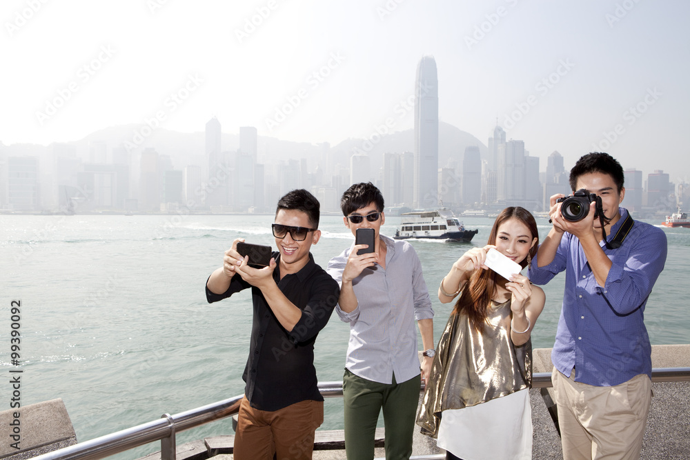 Fashionable young friends photographing with smart phones and SLR camera in Victoria Harbor, Hong Ko