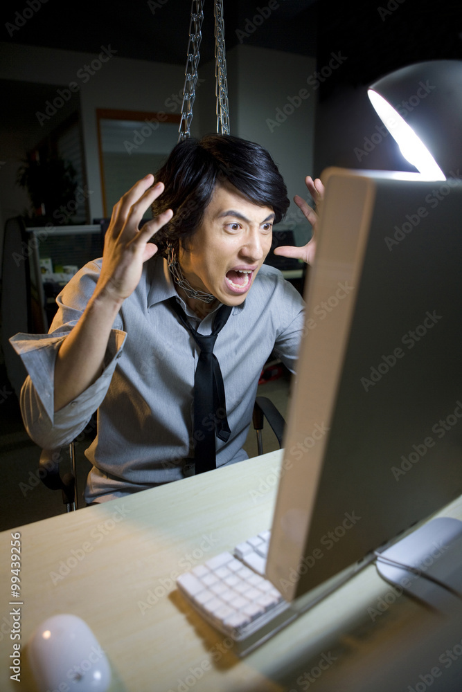 一个穿着衬衫打着领带的年轻愤怒男子在使用电脑时被挂在隔间的链条上