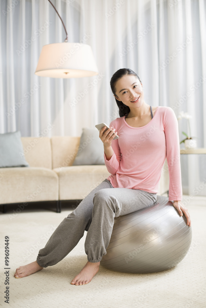 Young woman sitting on fitness ball with smart phone