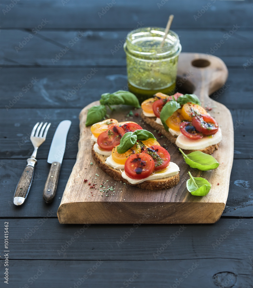 Tomato, mozzarella and basil sandwiches on dark wooden chopping board, pesto jar, dinnerware over bl