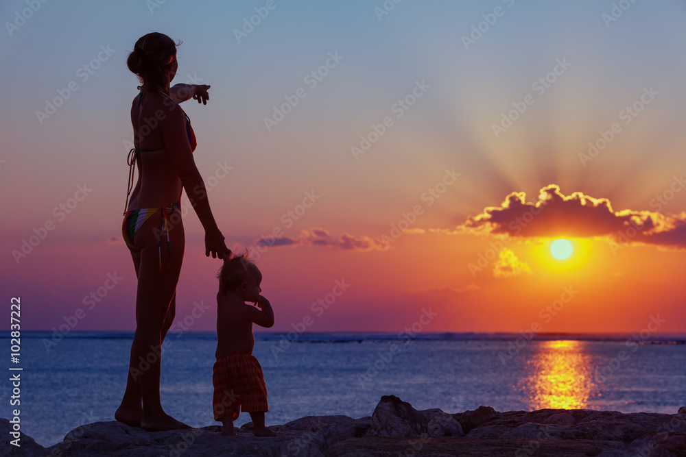 Black silhouette of happy family walking on sea beach - mother hold baby son hand, point sun and loo