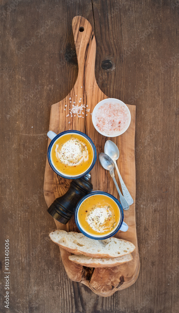 Homemade pumpkin cream soup in enamel mugs with herbs and fresh bread slices on olive serving board 