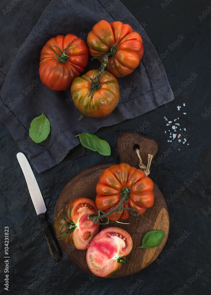 Fresh ripe hairloom tomatoes and basil leaves on rustic wooden board over black stone background.