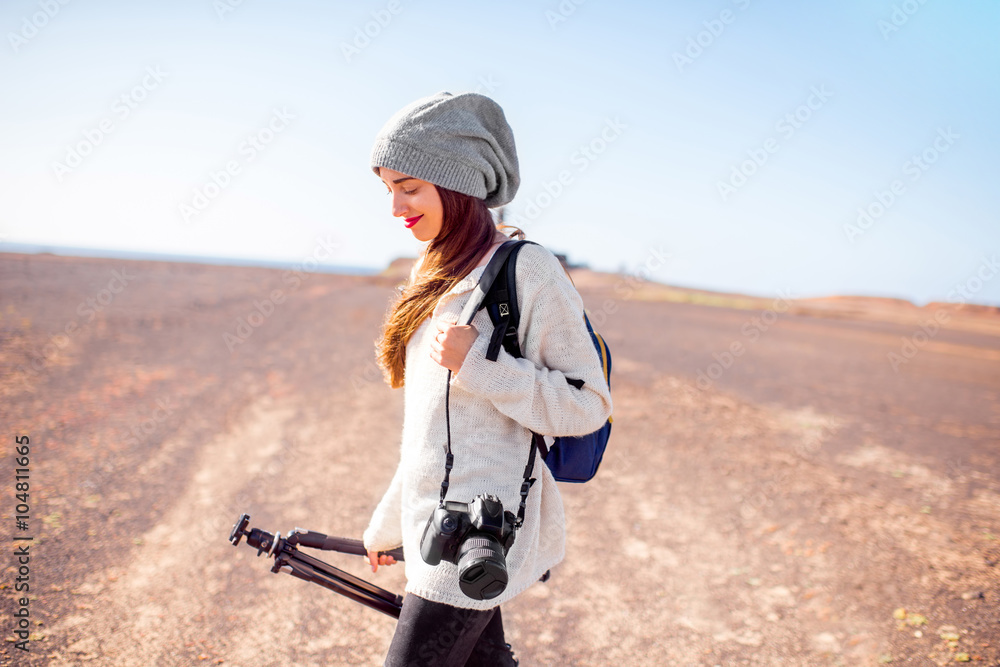 年轻的女摄影师穿着毛衣，戴着帽子，带着相机、背包和三脚架在沙漠中行走