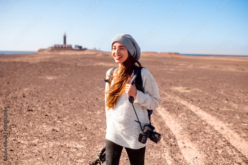 年轻的女摄影师穿着毛衣，戴着帽子，带着相机、背包和三脚架在沙漠中行走
