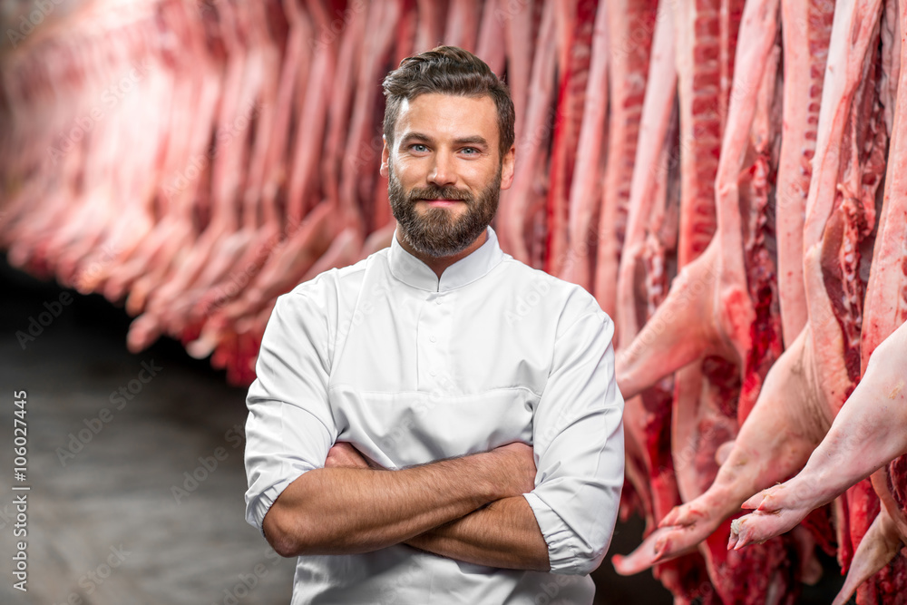 一位身穿白色制服的英俊屠夫在肉类制造厂的肖像，上面有猪肉尸体