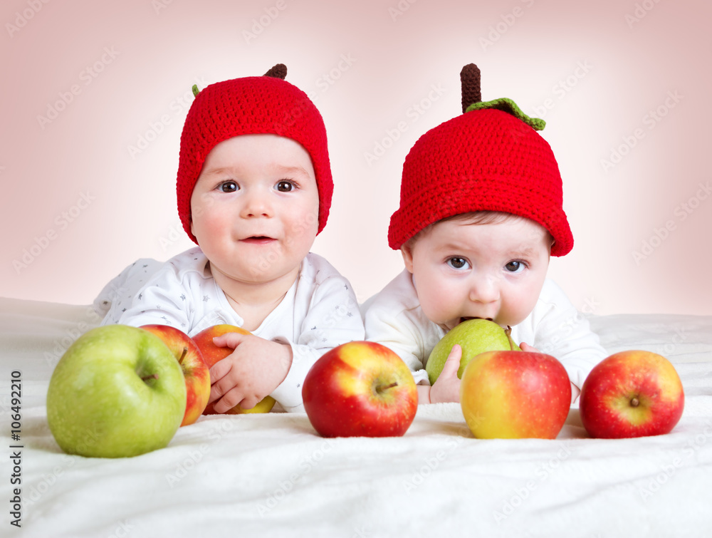 两个可爱的六个月大的婴儿戴着帽子躺在柔软的毯子上，上面放着苹果