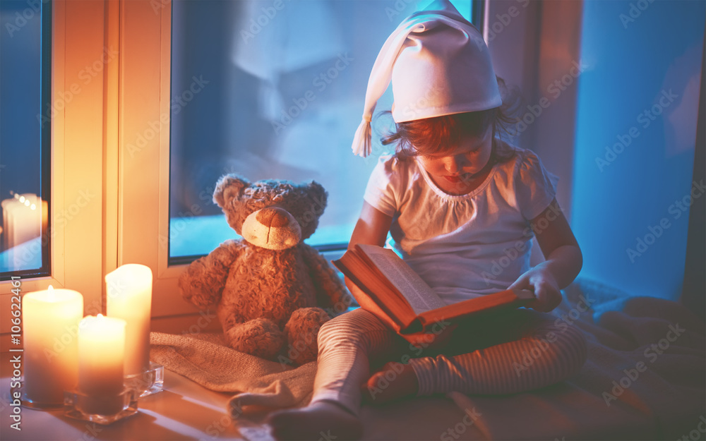 little child girl reading book by window before bedtime