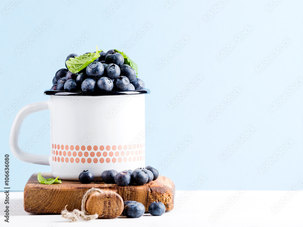 新鲜成熟的蓝莓，装在乡村风格的搪瓷马克杯上，装在蓝色柔和的背景上的乡村木板上