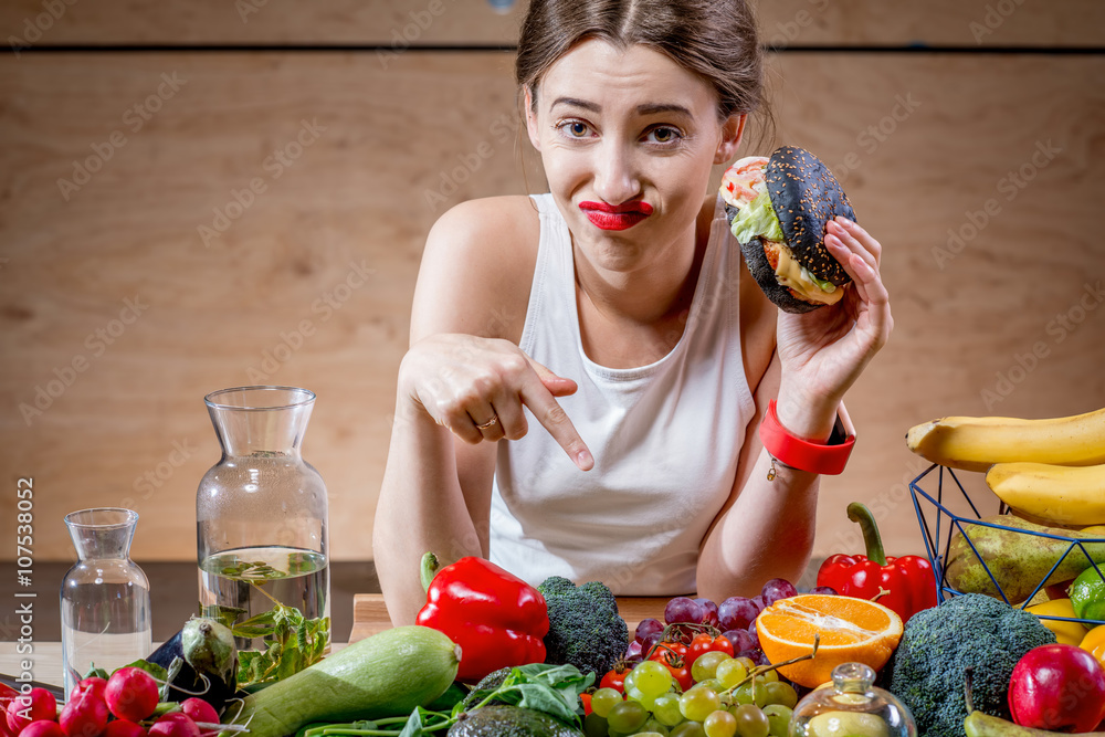年轻的运动女性在充满水果的餐桌上选择汉堡和健康但无味的食物