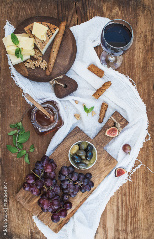Glass of red wine, cheese board, grapes, walnuts, olives, honey and bread sticks on rustic wooden ta