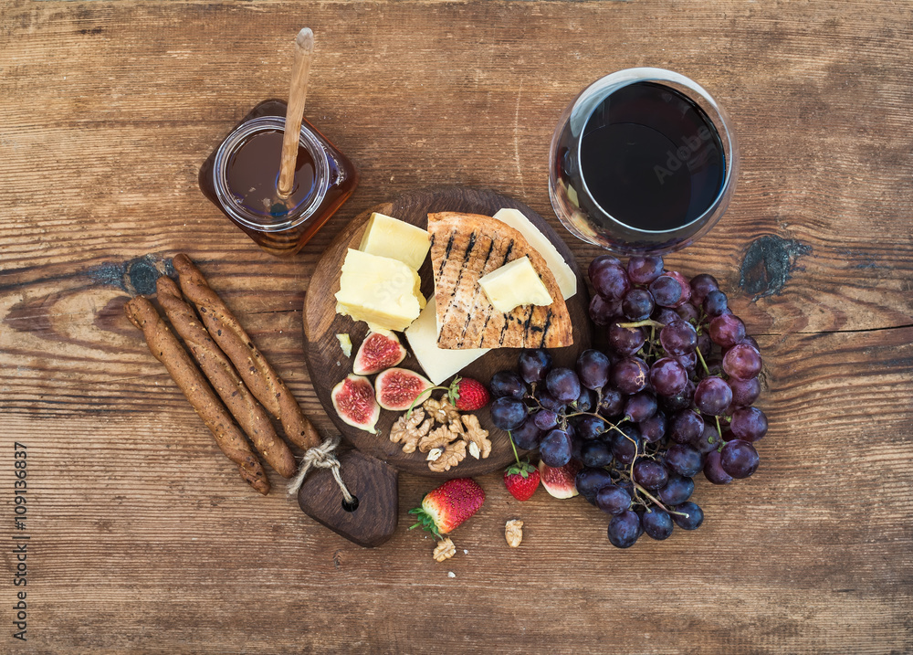 一杯红酒、奶酪板、葡萄、无花果、草莓、蜂蜜和面包棒放在乡村的木头上