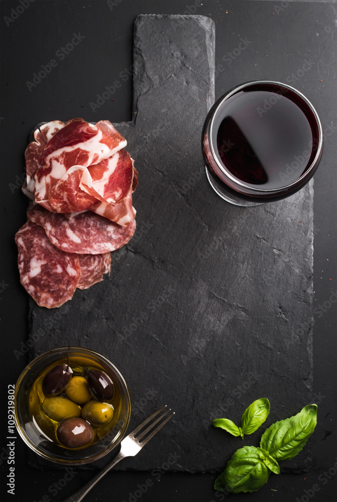 一杯红酒、肉类开胃菜、橄榄和罗勒，黑色石板石板，深色背景