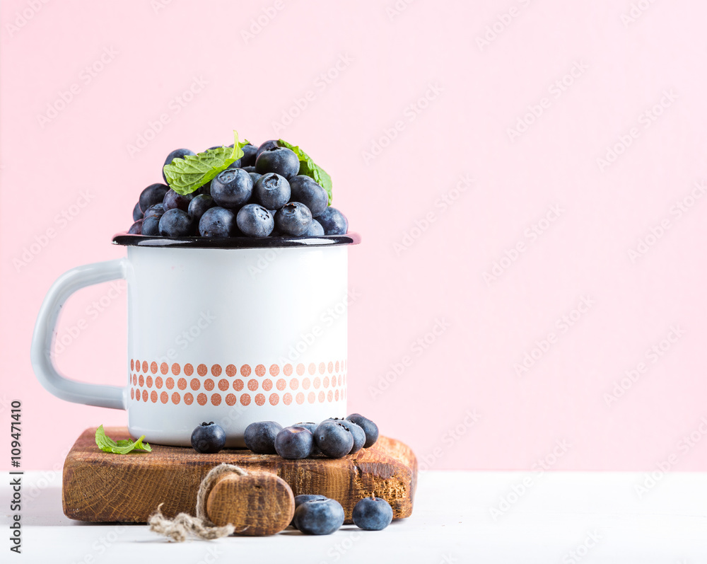 新鲜成熟的蓝莓，装在乡村风格的搪瓷马克杯上，装在柔和的粉红色背景上的乡村木板上