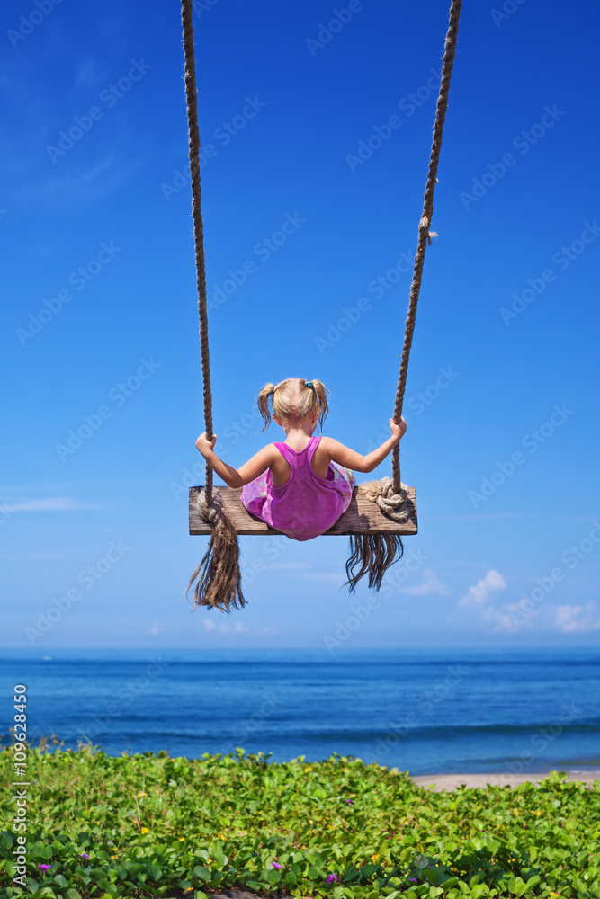 小女婴在蓝天背景下，在海浪和沙滩上，在绳索秋千上快乐地飞得很高。