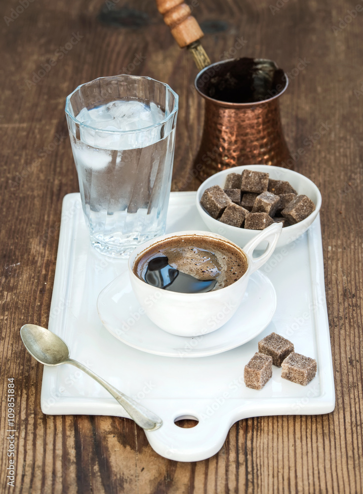 一杯黑咖啡，铜壶，玻璃杯里有冰块的水，白陶瓷餐具上有甘蔗糖块