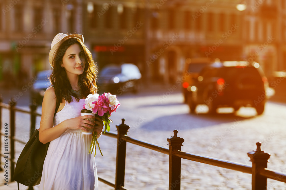 Portrait of young beautiful lady walking on street, holding paper cup of coffee & bouquet of peonies