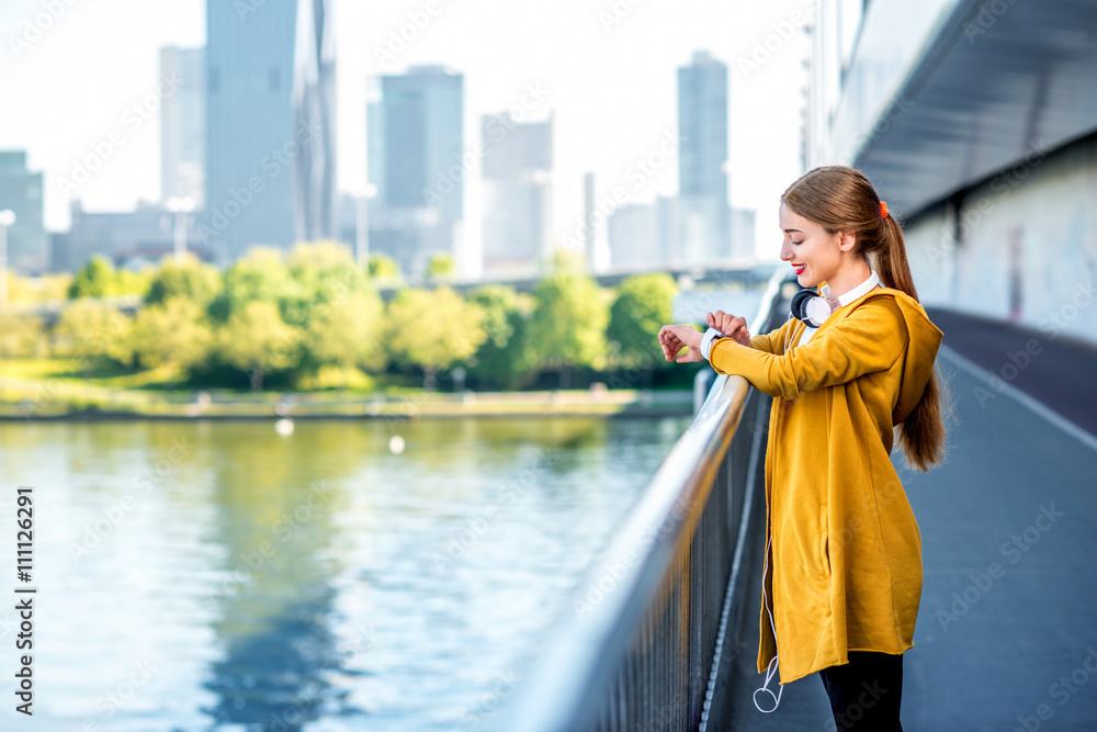 穿着黄色毛衣的年轻运动女性在摩天大楼林立的现代桥上看着智能手表