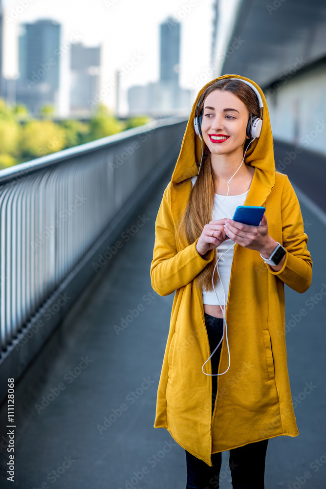 穿着黄色毛衣的年轻运动女性在现代桥牌上用智能手机听音乐