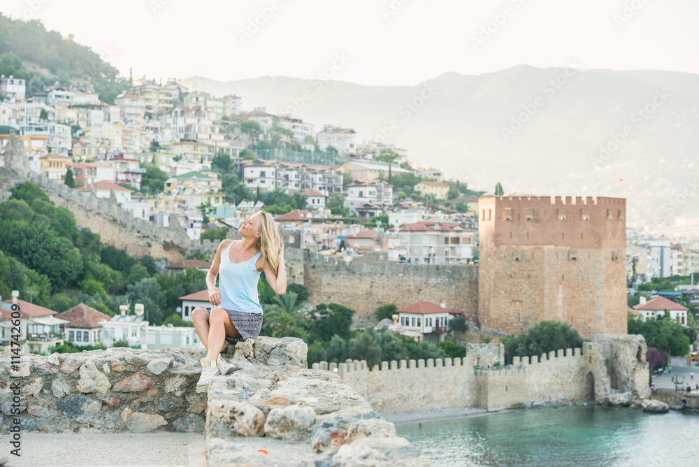 坐在阿拉尼亚城堡古老堡垒墙上的年轻金发女游客。