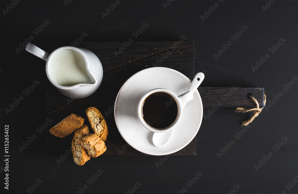 Cup of hot espresso, creamer with milk and cookies on dark rustic wooden board over black background