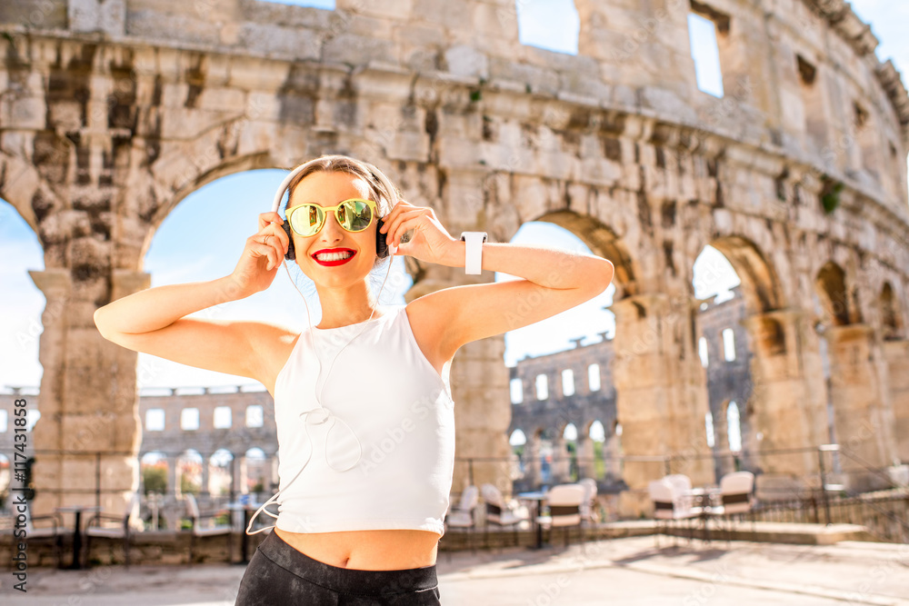 年轻微笑的运动女性戴着耳机站在古老的圆形大厅附近听音乐