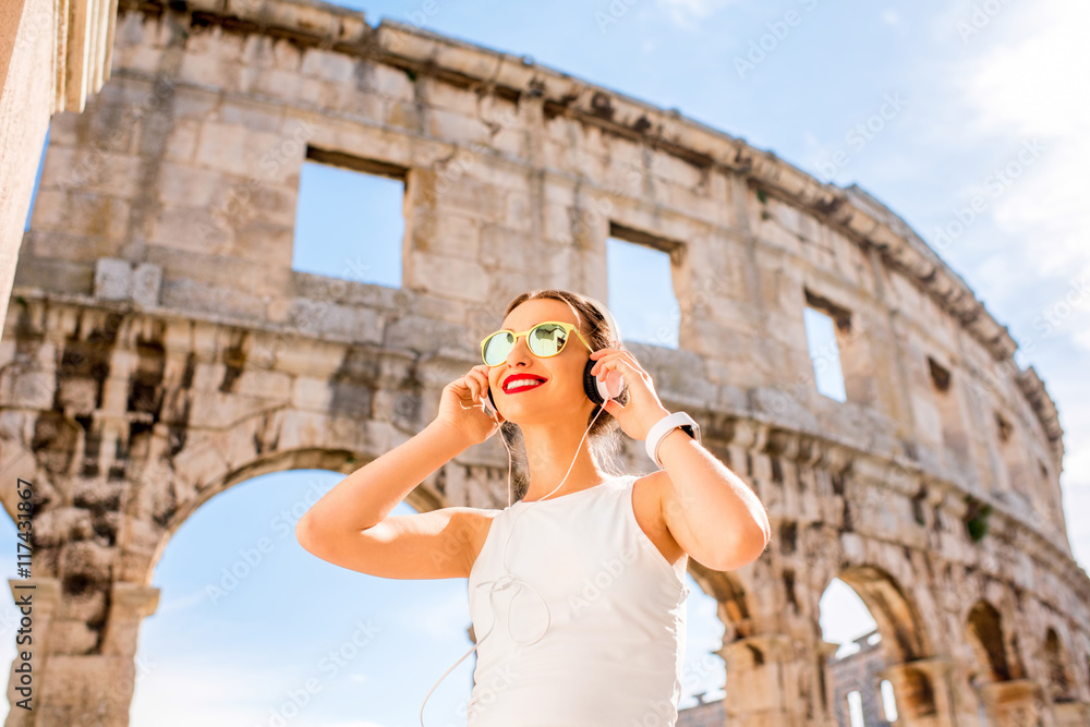年轻微笑的运动女性戴着耳机站在古老的圆形大厅附近听音乐