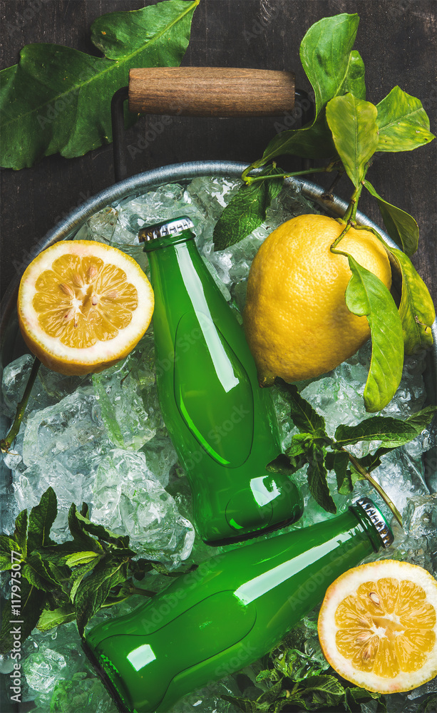 Bottles of green lemonade on chipped ice in metal tray with fresh lemons and mint over dark wooden b