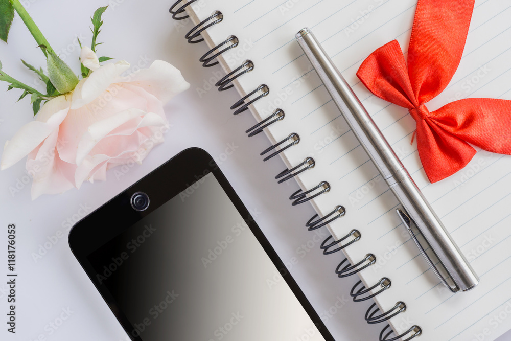 空白笔记本上的红色蝴蝶结和笔银，粉色玫瑰花和手机俯视图