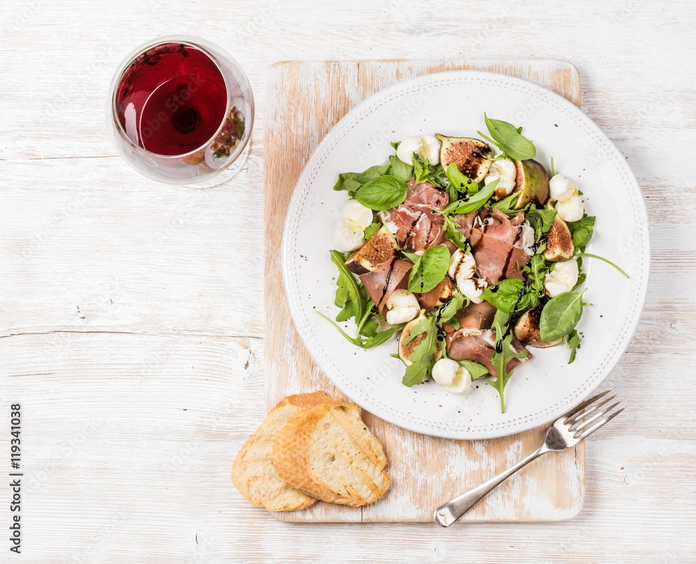 Prosciutto, arugula, basil, figs and mozzarella salad served with slices of bread on rustic board an