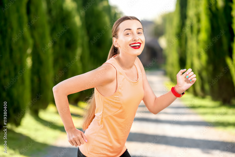 穿着橙色衬衫和黑色紧身裤的年轻运动女性在户外的小巷里与柏树一起奔跑。早上