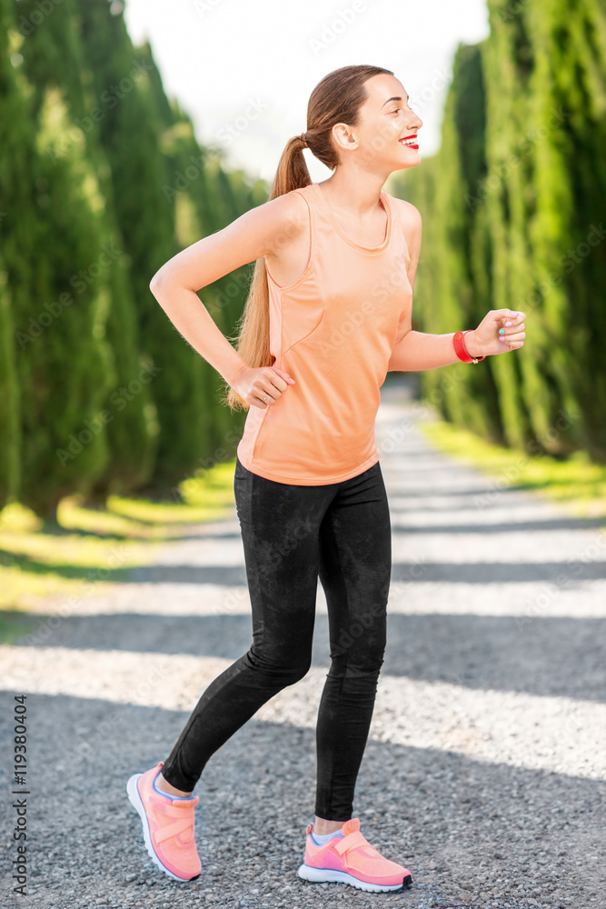 穿着橙色衬衫和黑色紧身裤的年轻运动女性在户外的小巷里与柏树一起奔跑。早上
