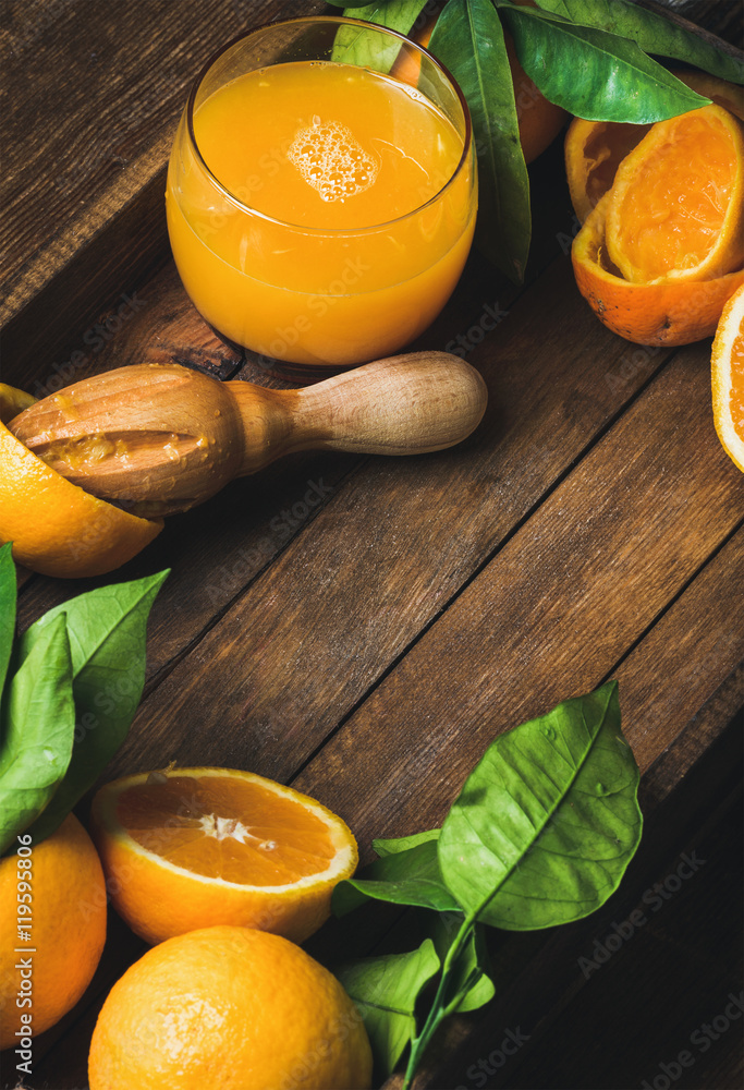 一杯鲜榨果汁和带叶子的新鲜橙子，放在深色木托盘里，放在木背上。