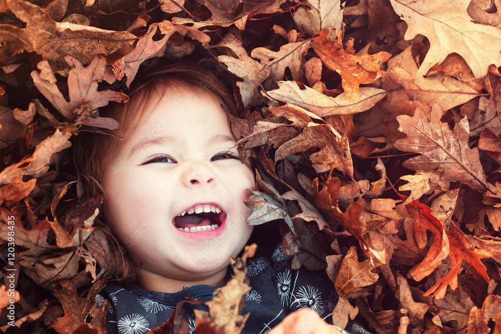 快乐的蹒跚学步的女孩躺在一堆树叶中微笑