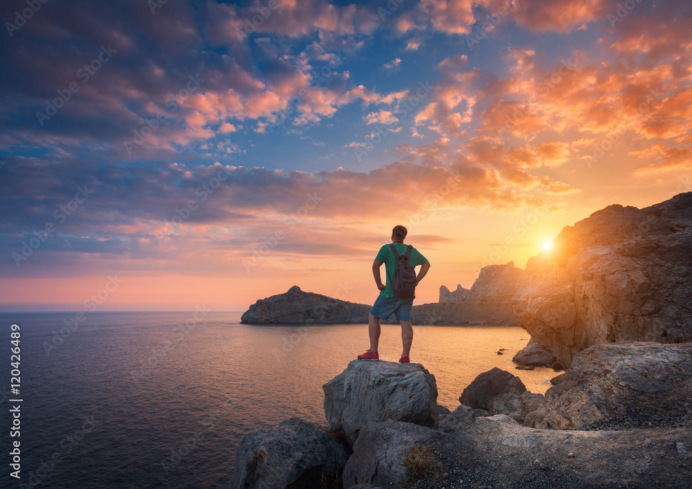 Young standing man with backpack on the stone on the seashore at colorful sunset sky. Beautiful land