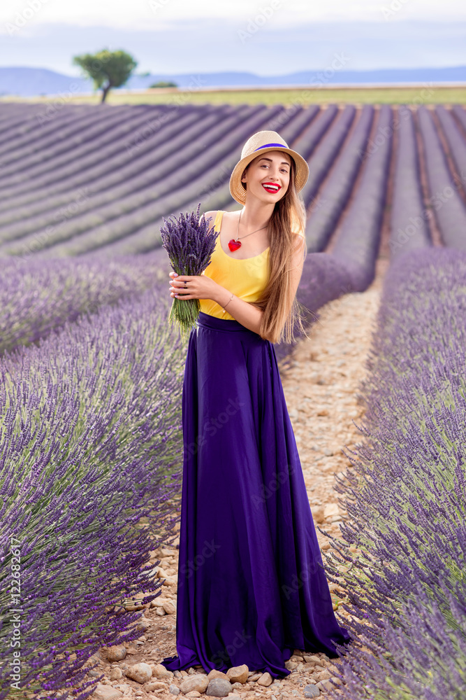 年，一位身穿紫色长裙的年轻美女手持薰衣草花束站在薰衣草地上