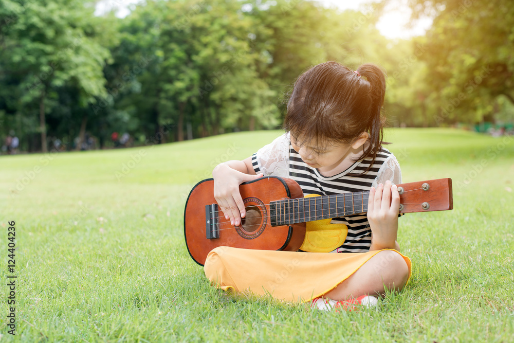 亚洲小女孩坐在草地上，在公园里玩尤克里里琴