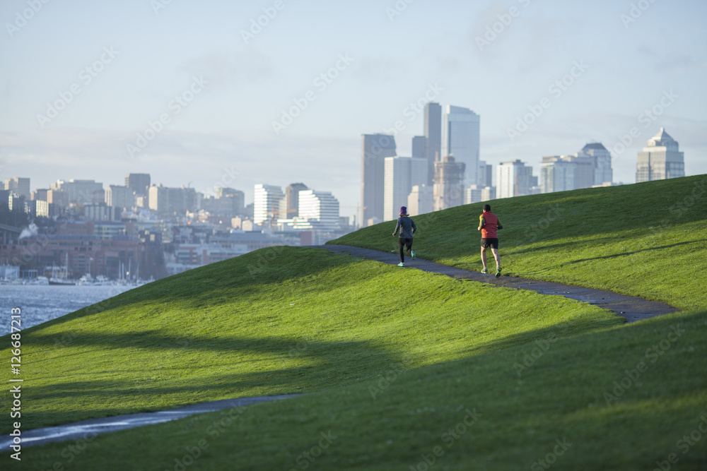 一对成年男女在穿过煤气厂公园的铺好的小路上奔跑，西雅图的城市景观就在眼前