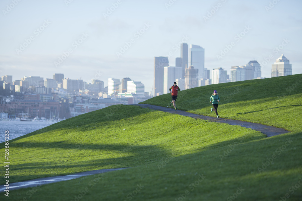 一对成年男女在穿过煤气厂公园的铺好的小路上奔跑，西雅图的城市景观尽收眼底