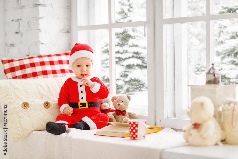 快乐的婴儿打扮成圣诞老人坐在家里的窗户上