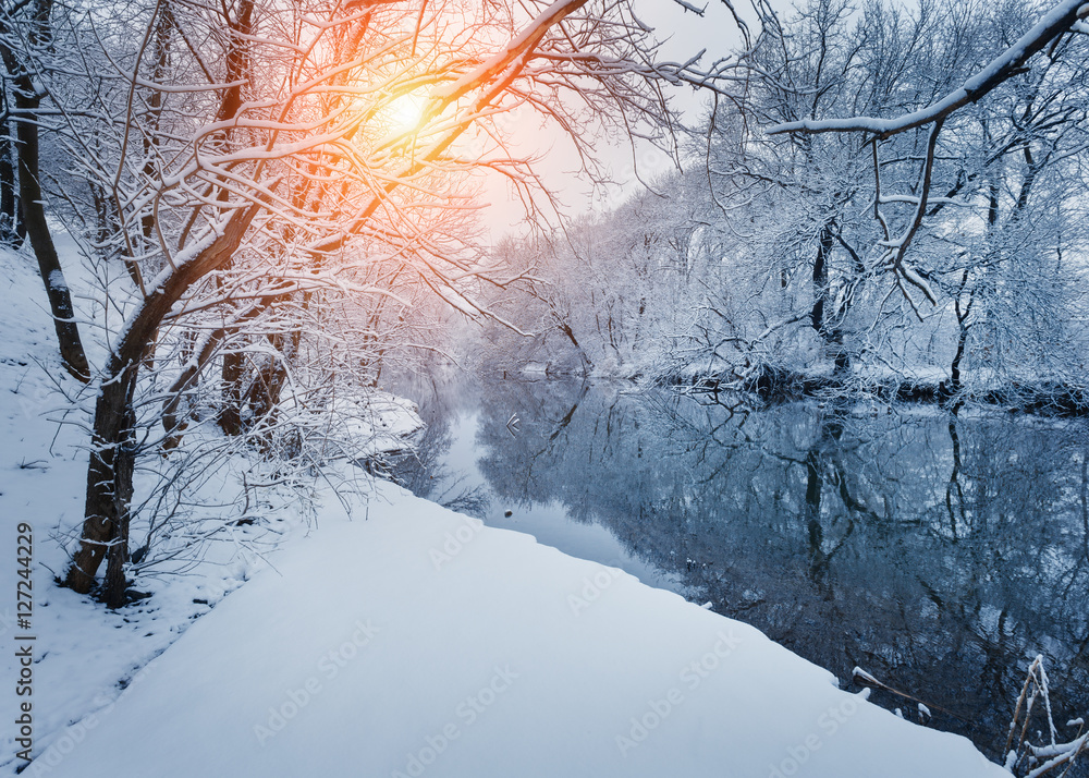 日落时河边的冬季森林。白雪皑皑的树木点缀着五颜六色的景观，冰冻的河流倒影着